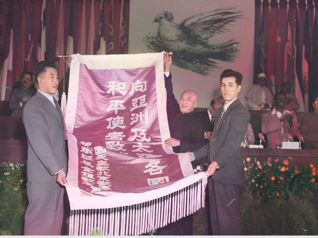 1952年10月北京“亚洲及太平洋区域和平会议”会场上，马共代表连克苏（右）代表与会和平使者接受中国天主教北京教区神职班及全体教友敬献的“向亚洲及太平洋各国和平使者致敬”锦旗