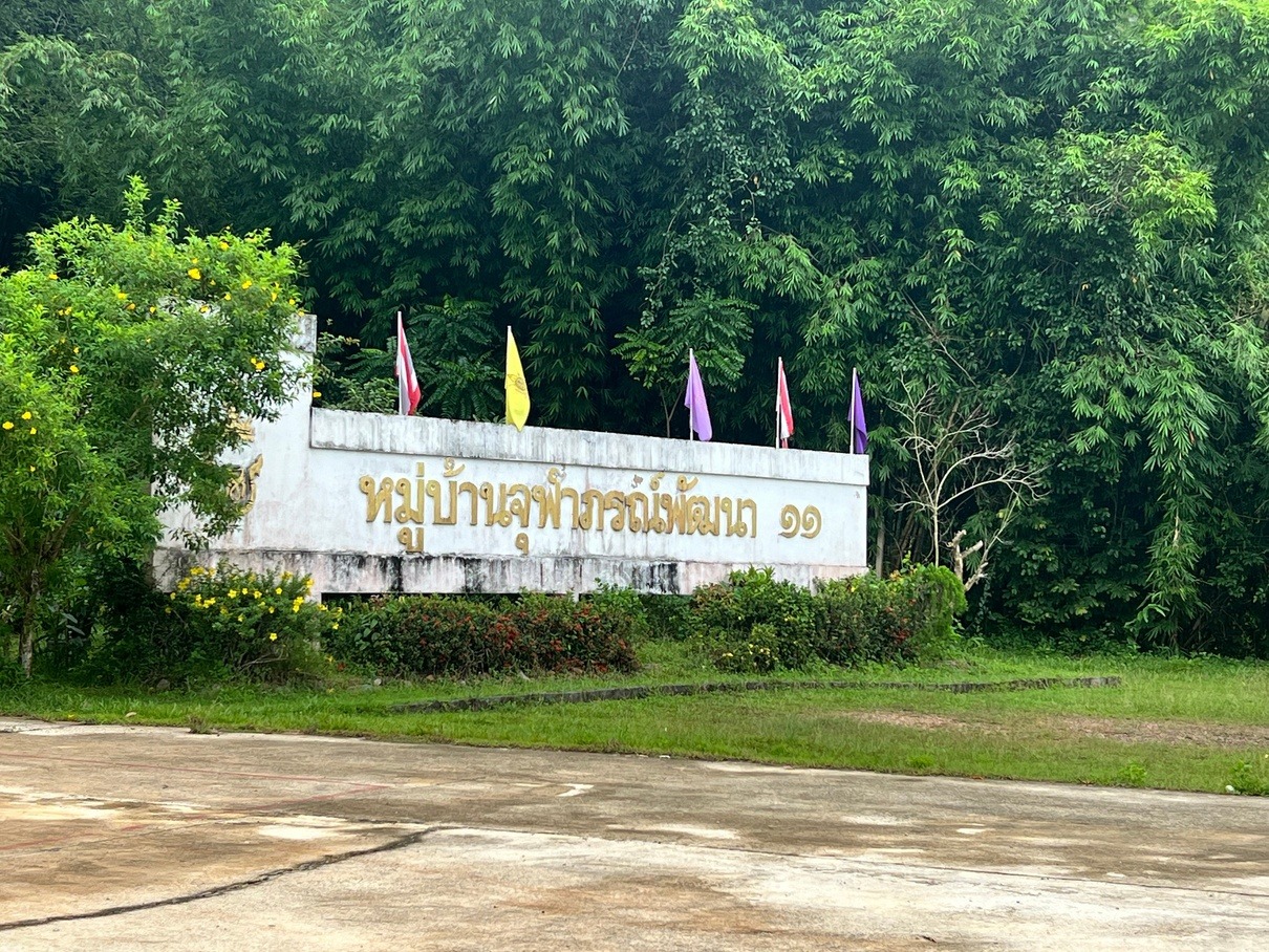也哈和平村（第11朱拉蓬公主发展村）庄重的泰文村名石碑（หมู่บ้านจุฬาภรณ์พัฒนา ๑๑），碑后彩旗与泰国国旗迎风飘扬。