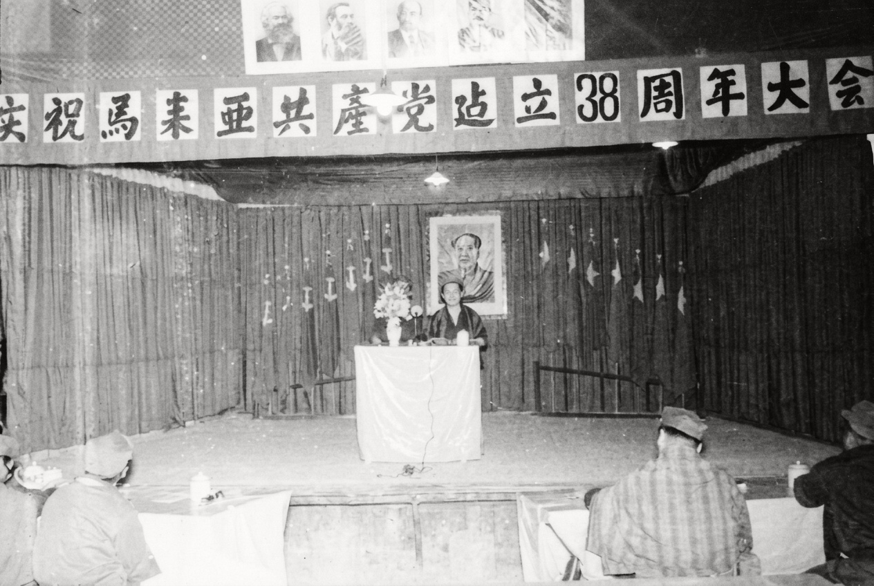 1968年密林根据地举行的“庆祝马来亚共产党成立38周年大会”主席台，横幅上方悬挂马恩列斯毛画像，台后并列十四面马共党旗与民族解放军军旗。