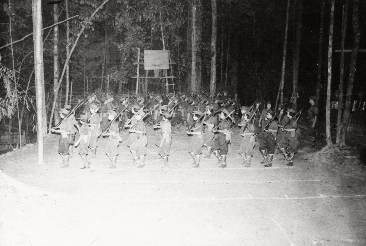 1968年6月20日庆祝抗英民族解放战争20周年升旗礼上，肩扛步枪、裹扎绑腿的马来亚民族解放军纵队在热带雨林营地列队行进。